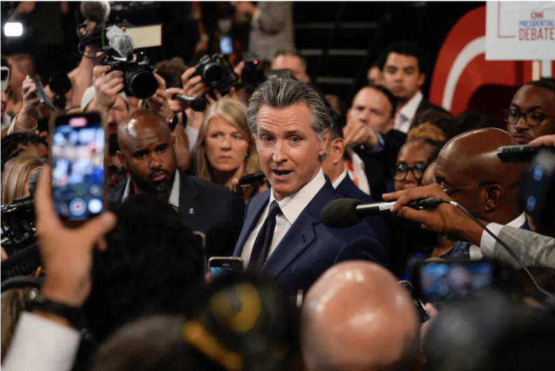 Gavin Newsom, dressed in a suit, is in the middle of a crowd of reporters, many taking pictures and recording with their phones.