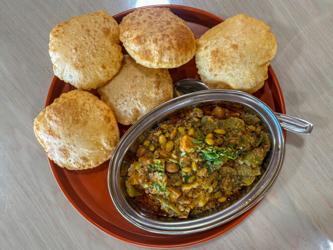 An overhead shot of the Undhiyu Puri at Jay Bharat — a dark, complex Gujarati vegetable curry with visible legumes and a fresh herb garnish in a silver kadai, surrounded by five golden puffed puris on a red tray