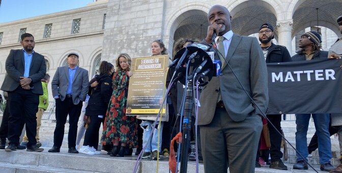 LA City Council Member Marqueece Harris-Dawson wears a grey suit with beige tie and holds a microphone. LA City hall can be seen in the background. He is surrounded by activists. Some of them hold a Black Lives Matter sign.