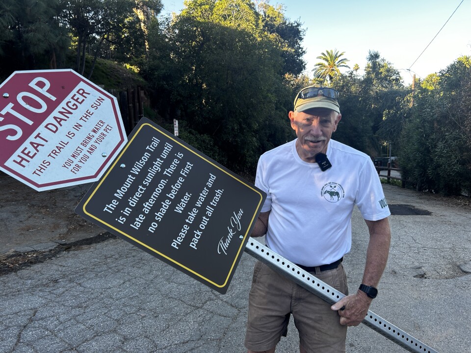 An older white man wearing a white t-shirt and baseball cap and tan shorts holds a large sign that says "stop heat danger" among other things. 