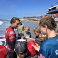 A wetsuit-clad surfer signs hats and a surfboard for a group of children gathered on the beach near a competition venue. One of the kids has a shirt that reads "Colapinto" on the back. 