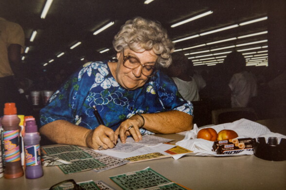 A close up of a light skinned woman with gray hair, glasses and a blue dress at a table playing bingo. She has Snickers bars and peaches by her side.