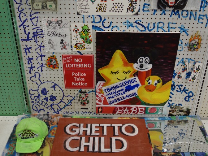 A small display of several different artworks and objects on a converted supermarket shelf. One item reads "ghetto child" while another reads "No loitering, police take notice."