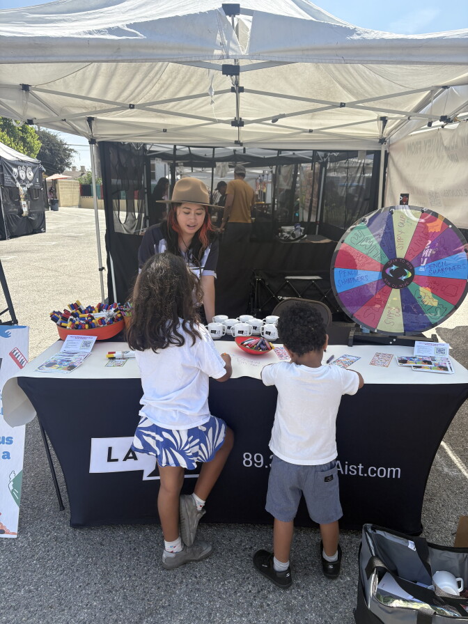 An outdoor photograph captures a community booth hosted by LAist, set up under a white canopy tent on an asphalt surface. A staff member wearing a brown wide-brimmed hat stands behind a table draped with a black tablecloth that features the LAist logo and text reading "89.3 LAist.com". Two young children with curly dark hair, viewed from behind, stand at the front of the table looking at the booth's activities. On the table sit white mugs, a red bowl filled with markers, informational flyers, and an orange basket holding pens. To the right, a large colorful prize wheel is positioned for a game, displaying sections labeled with items like "sippy cup" and "pencil sharpeners".