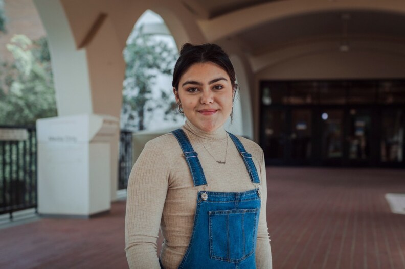 A young woman with light brown skin smiles at the camera. Her dark hair is parted in the middle and pulled back and she wears a beige turtleneck and denim overalls. She stands on the large brick patio of an academic building. Stone arches can be seen behind her.