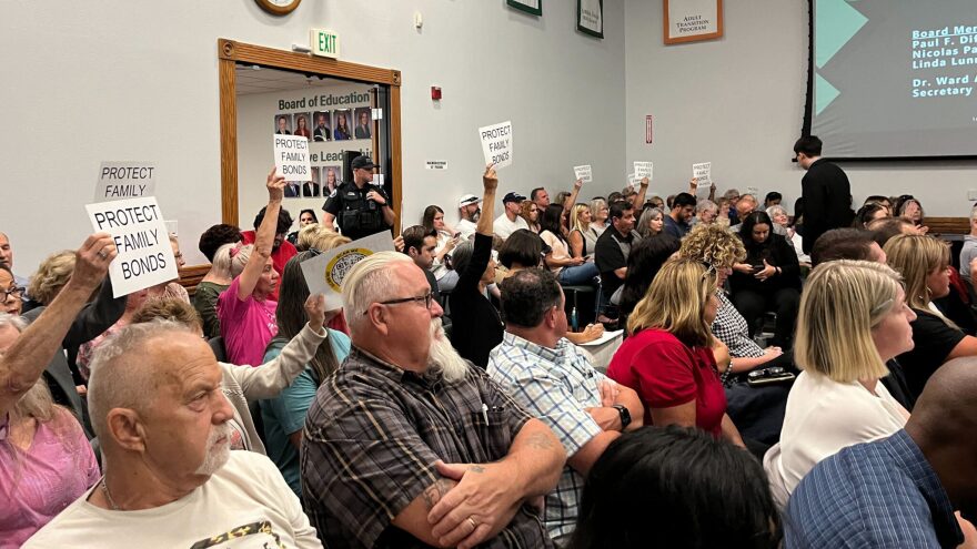 A crowd of people seated in chairs in a large room. They face the same way, many holding signs which say PROTECT FAMILY BONDS. A male-presenting figure in the center crosses their tattooed arms. A police officer stands in the background in a wide wooden doorway.