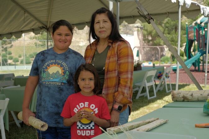 An older woman with dark hair stands with her young grandchildren outside under a shade structure. 