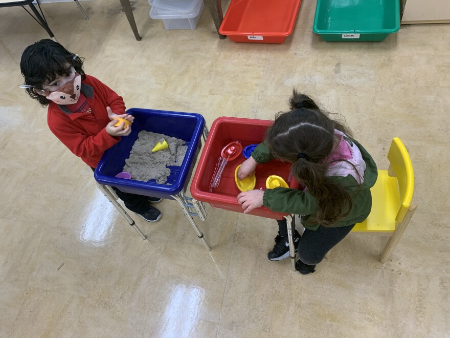 Two children play in a classroom. A girl with pigtails reaches her hands into a tub of water and yellow plastic boats. A boy with a red shirt squishes sand in a blue tub.