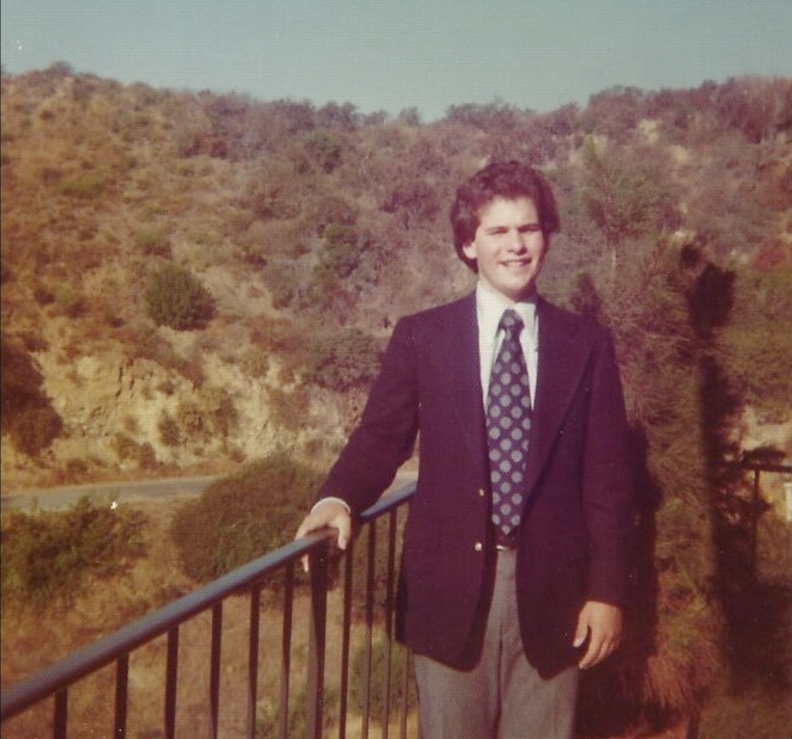 A young man in a suit jacket and tie has curly hair and light-tone skin. He poses at a railing with a view of brush-covered hills behind him.