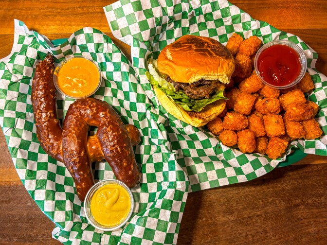 A meal spread featuring a cheeseburger with lettuce and tomato, golden tater tots with ketchup, and two soft pretzels served with mustard and cheese sauce, all on green checkered paper.