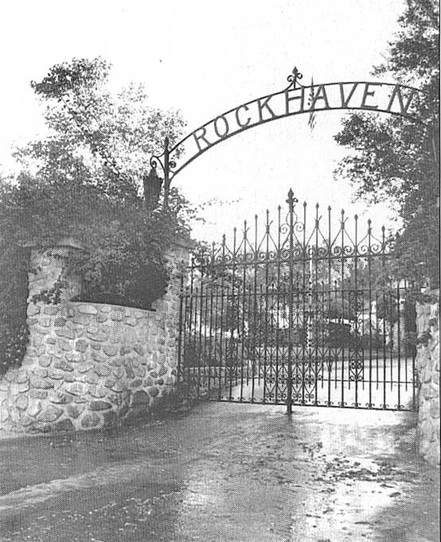The front metal gate, made of vertical bars, of Rockhaven Sanitarium, with the word "Rockhaven" in an arch above the gate. A house is visible in the background through the gate's bars.