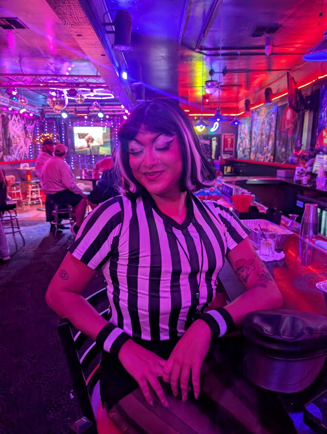 A drag performer wearing a soccer jersey sits at a bar, smiling.