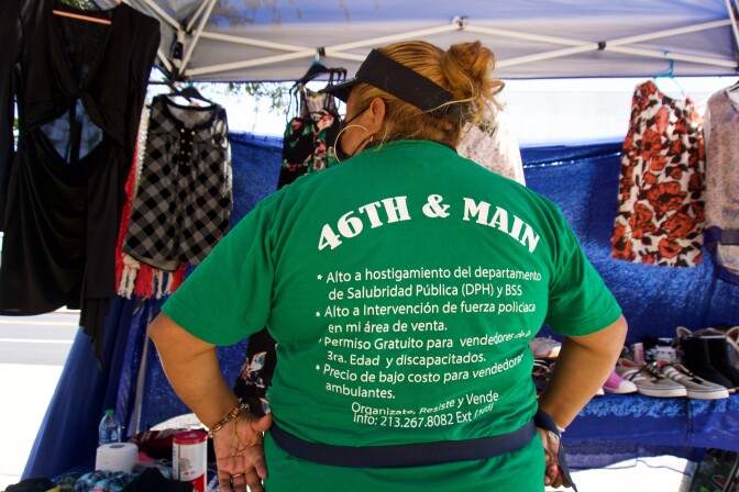 a woman wearing a face mask stands with her back to the camera. her shirt reads "46th & Main" and then lists street vendor demands in Spanish