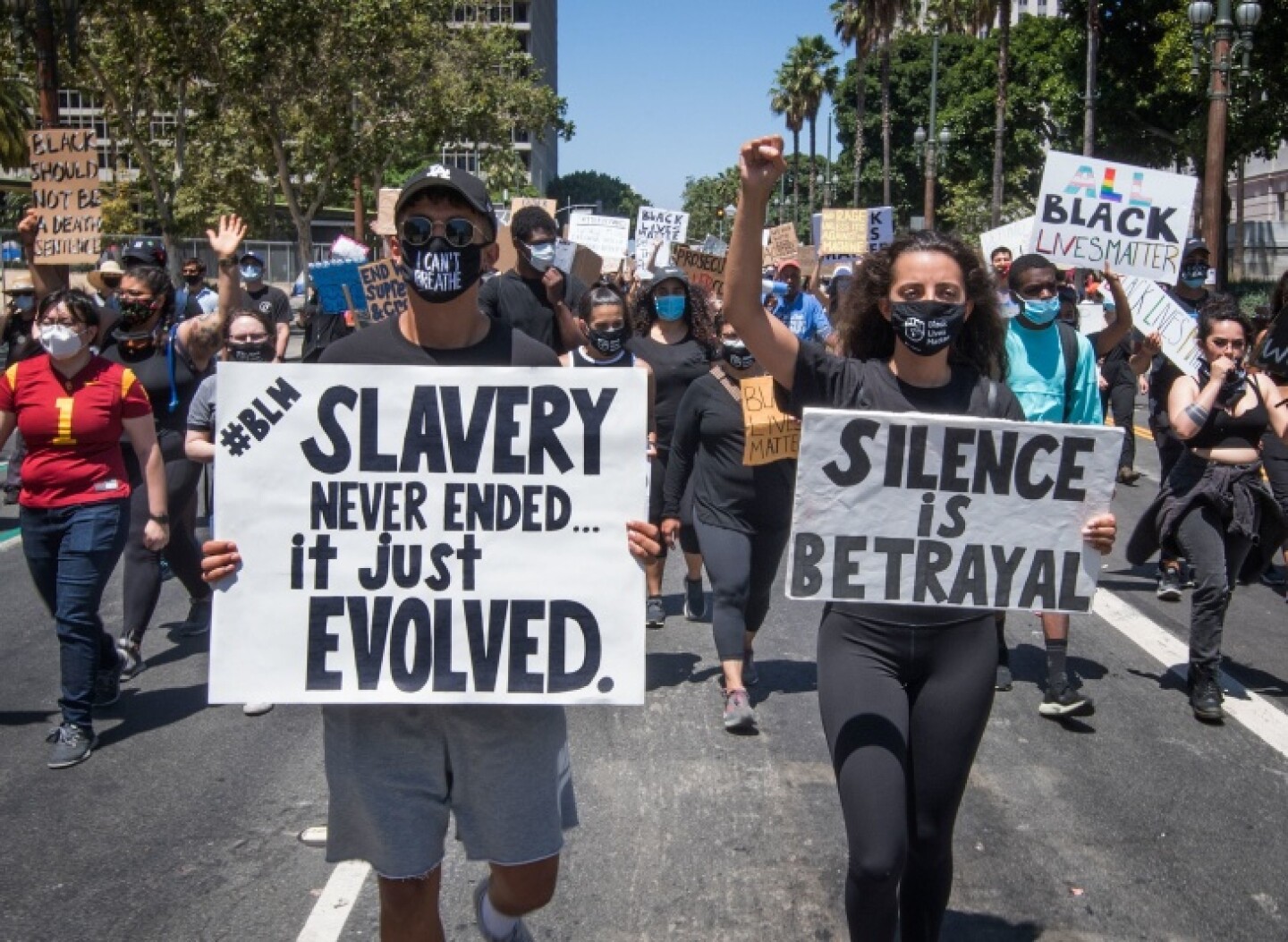 A man wearing a mask that says I Can't Breathe and carrying a sign that reads "#BLM Slavery Never Ended.. It just evolved" walks beside a woman wearing a mask and carrying a sign that reads "Silence is Betrayal" during a protest in downtown L.A. Dozens of people are walking behind them carrying protest signs.