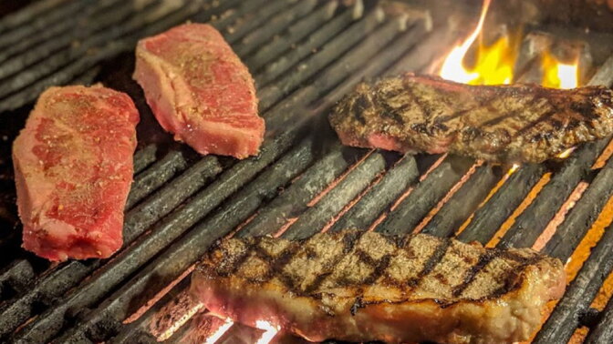 Four raw and grilled steaks cooking on a flaming charcoal grill, two seared with grill marks and two still raw.