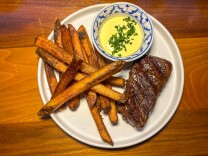 A neatly plated steak frites dish featuring a seared, medium-rare cut of steak with visible grill marks, thick-cut golden fries, and a small bowl of creamy yellow sauce garnished with chopped chives, all served on a white plate atop a warm wooden table.