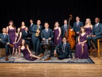 A group of 16 people people all posing. From left to right: A light-skinned man wears a navy blue suit and purple tie with brown shoes and sits on a chair with legs crossed. To the right, a light-skinned woman stands next to him wearing a purple dress. Another light-skinned woman wearing a purple dress is sitting next to her. A light-skinned woman is partially laying down on a blue and white rug also wearing a purple dress. To her right, a light-skinned man wearing a navy suit sits on a chair and places hands on a drum. To the right, a medium-light toned man sits on the rug wearing a navy suit. To his right, a light-skinned woman wears a purple dress and sits on a chair. To her right, a dark-skin toned man sits on top of a tall chair. To his left, a light-skinned woman wearing a purple dress. To her left, a light-skinned man wears a navy suit. To his left, a light-skinned man wearing a navy suit holds a cello. To his left, a light-skinned woman with blonde hair wears a purple dress. To her left, behind the man with the drum, a light-skinned man wearing a navy suit holds an alto-saxophone. To his back left, a medium-light skin toned man stands wearing a navy suit. In front, a light-skinned toned man wearing a navy suit sits on a chair. Behind him, a light-skin toned woman stands wearing a purple dress. 