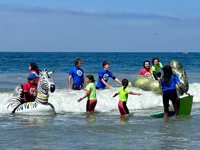 A small group of young adults and children stand in the shallow water wearing blue, neon green, and red shirts over wetsuits. One person is sitting on a unicorn-shaped raft and another is on a dinosaur-shaped raft. 