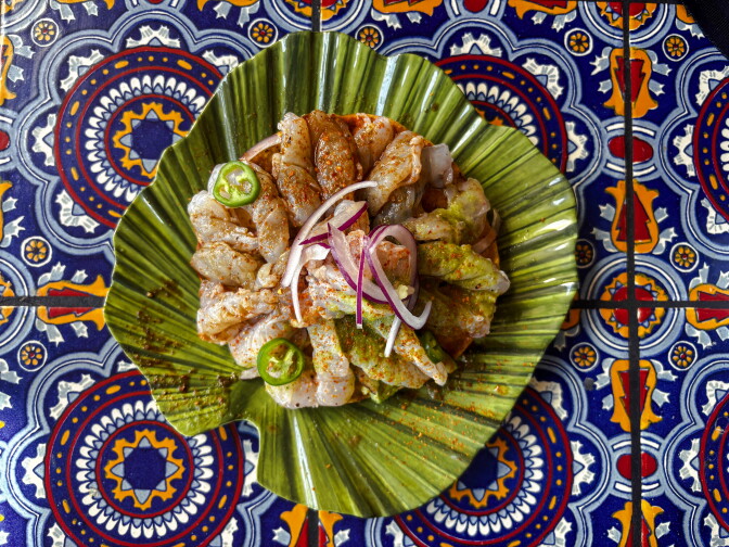 Shrimp aguachile topped with sliced red onion and jalapeño, served on a banana leaf–shaped ceramic plate atop a blue Talavera-tiled table.