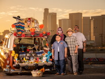 A man, a woman and two children stand next to a vintage station wagon decorated with Dia de los Muertos paraphernalia. In the background is downtown L.A. The sun is setting.