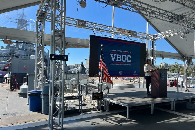 A female presenting person in slacks and a white shirt speaks at a podium in front of a blue screen with the letters VBOC. An American flag is visible on the stage. Scaffolding as well as a battle ship is visible in the background. 
