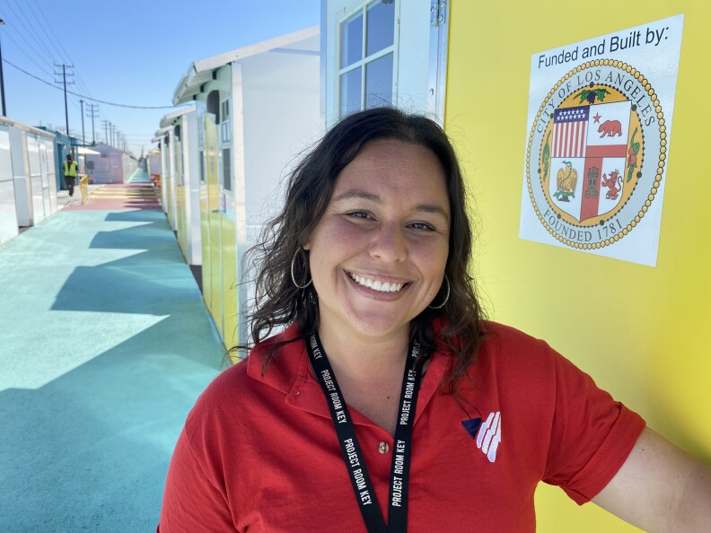 Desiree Martin, dressed in a red shirt and smiling, stands in front of a row of tiny homes at Branford Village in the Sun Valley.