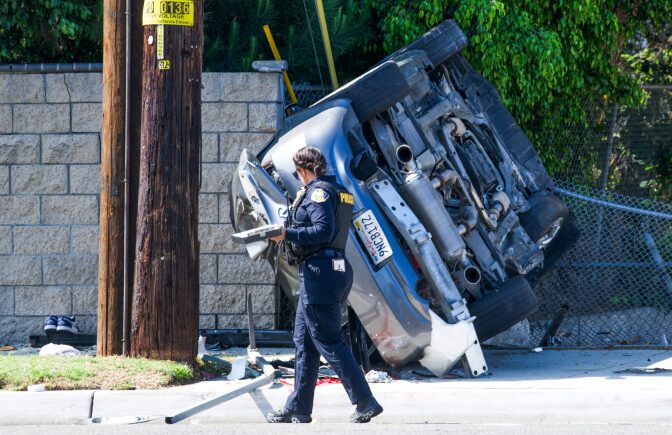 A uniformed police officer walks past a silver sedan that is on it's side, resting against a hollow block fence. There is a wooden electrical pole to the left of the vehicle.