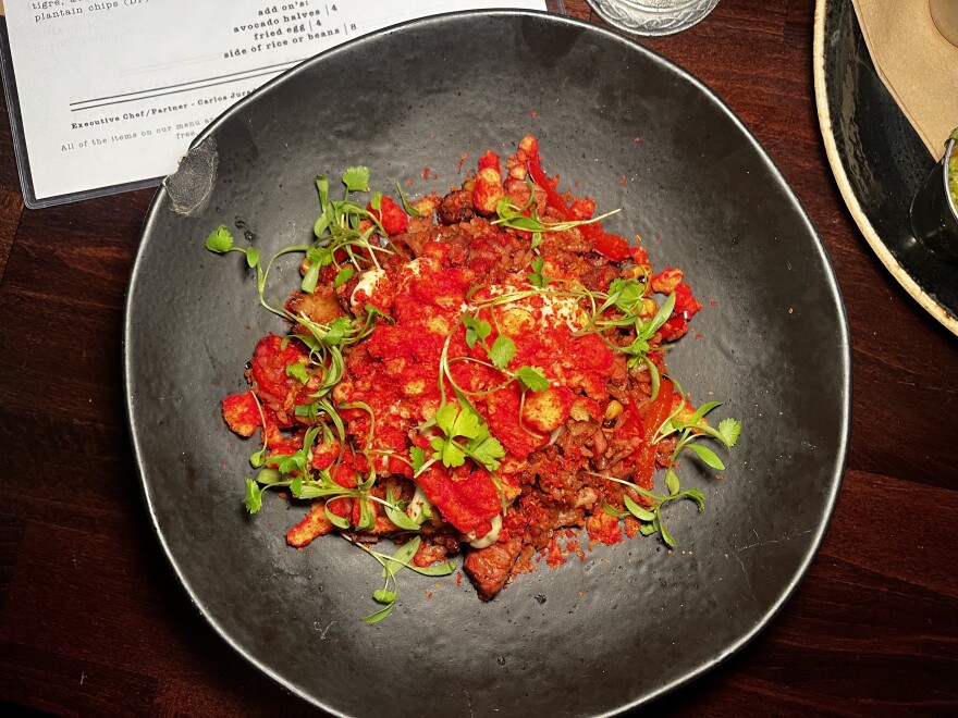 In this table setting, there is a serving of fried rice that has been garnished with a generous amount of crushed Flamin' Hot Cheetos. The Cheetos are a bright red color and can be seen in both their standard form and in smaller, crushed pieces. The dish is further adorned with a variety of light green herbs that add a refreshing touch to the overall presentation. The round plate itself is a subtle shade of grey, providing a neutral background that allows the vibrant colors of the food to pop.