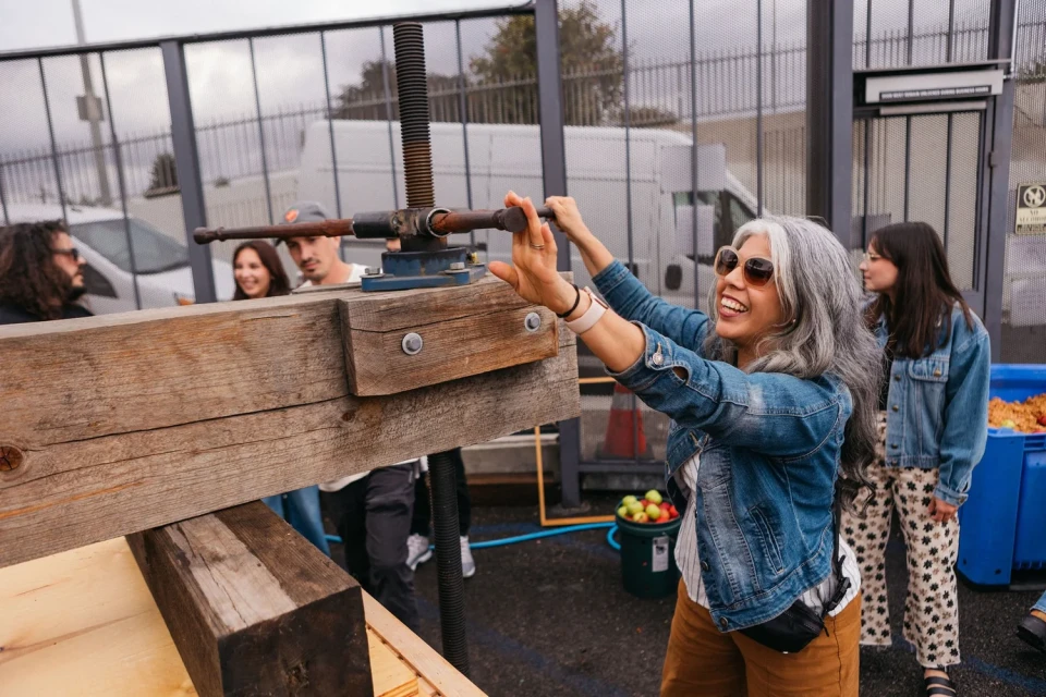 a woman with medium skin tone wears a denim jacket and turns an apple press while four people watch in the background