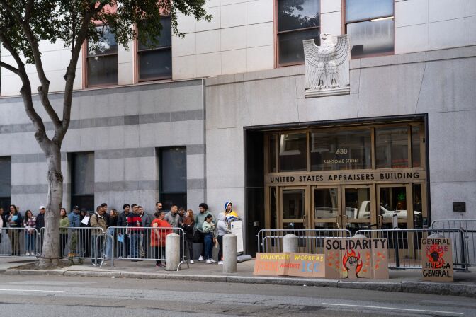 People stand in line behind a gated fence alongside a building, which has a bald eagle statute above the entrance and signage that reads "United States Appraisers Building." There are handmade cardboard signs with text that reads "Unionized workers strike against ICE," "Solidarity" with a fist on fire, and "General strike. Huegla General" with a fist on fire.