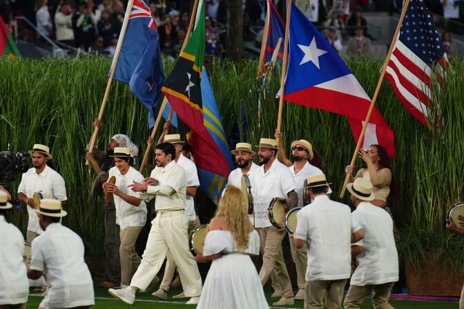 Bad Bunny, a man with medium skin tone, wearing a cream cream, pants, and shoes, sings as he walks leading a group of people holding various flags past an area of tall grass and other performers holding instruments.