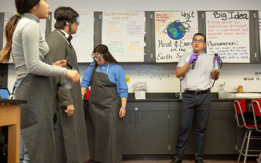 Three students wearing goggles and lab aprons stand next to their teacher who is talking into a microphone while holding a glass beaker.