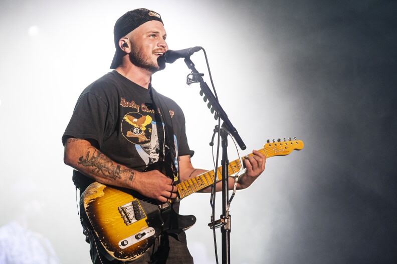 Country musician Zach Bryan perform onstage under a spotlight. He's playing an electric guitar and singing as he looks out into the crowd.