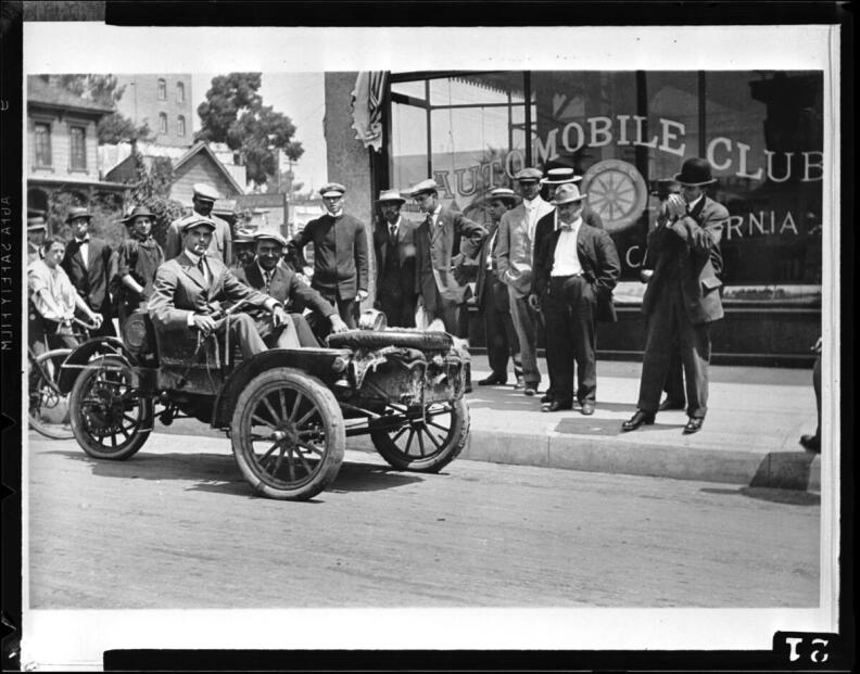 A black and white photo from 1912 shows two men in an early automobile as several other men look on in front of a large glass window with the words Automobile Club written on it.