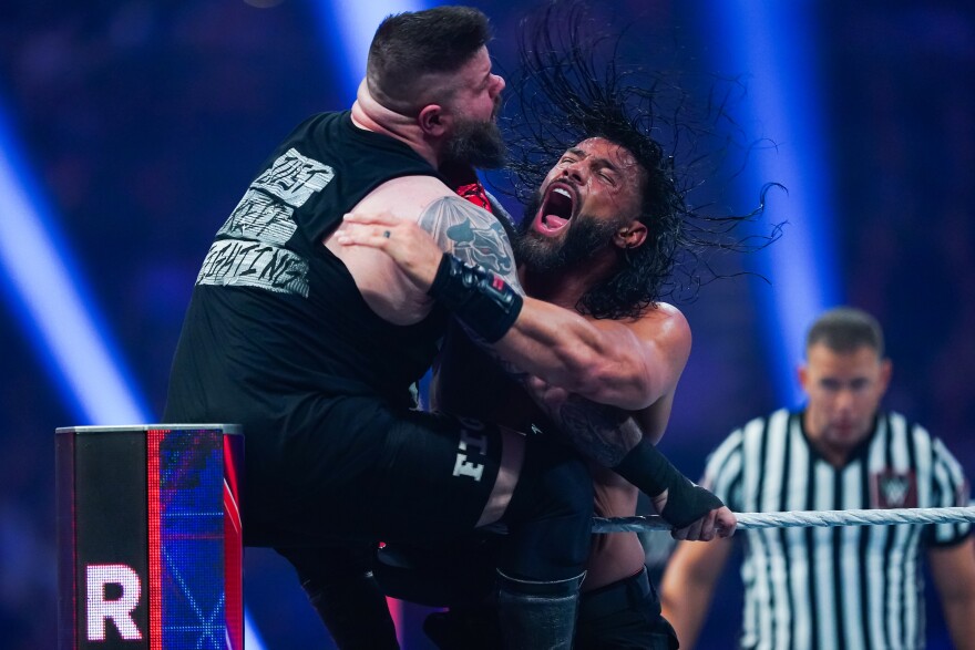 Roman Reigns' hair flies as he performs a wrestling move against wrestler Kevin Owens as Owens sits atop the ring post in the corner of a professional wrestling ring. A referee in a black and white vertically striped shirt looks on in the background. Reigns is Samoan, while Owens is white.