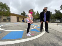 A man in a navy suit and purple tie bends down to chat with a young girl wearing a shiny pink coat. They are on a cement surface outside a one-story school building, and both are wearing masks.