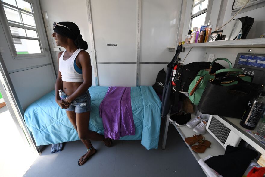 A woman stands inside a so-called tiny home, which includes a bed, shelves and storage cabinets. She is wearing a white tank top, blue jean shorts and sandals as she looks out her open door.