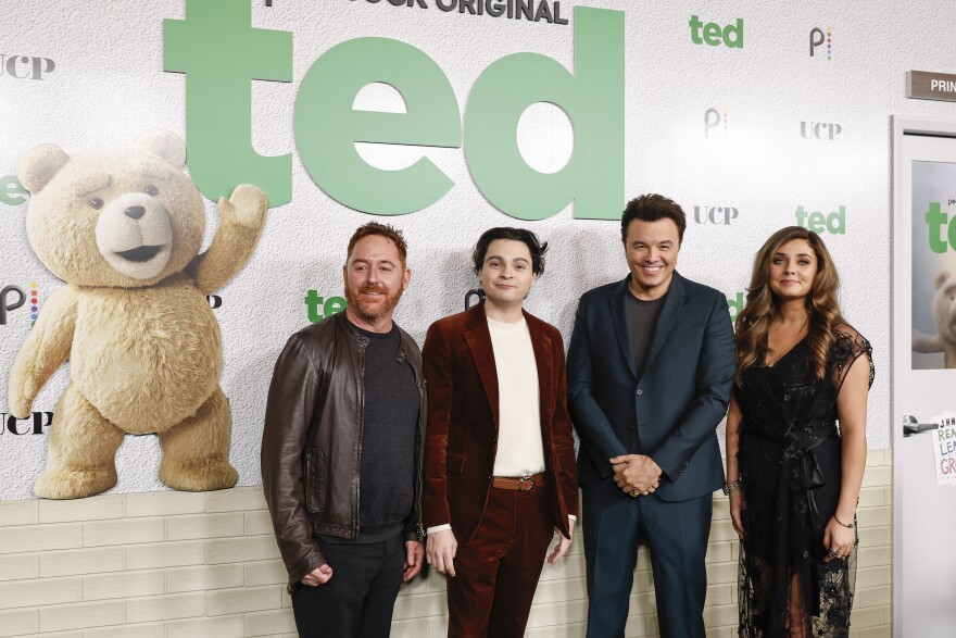 Three men and a woman stand side-by-side in front of a large backdrop featuring a large anthropomorphic Teddy bear next to the word 'Ted'