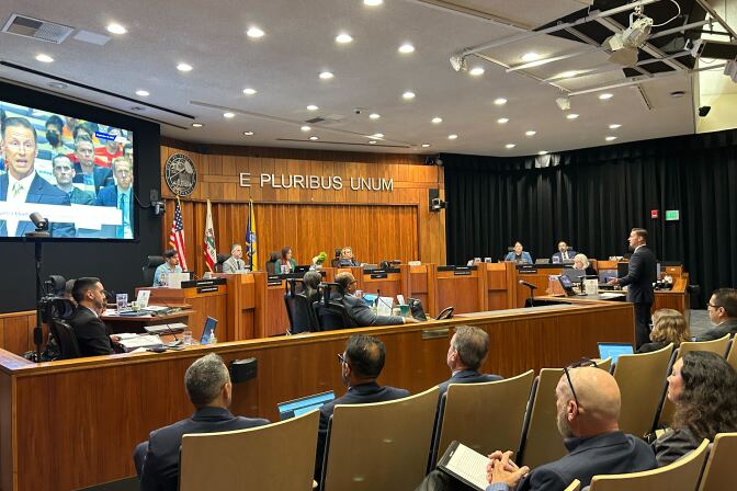 A row of people are seated behind a panel with a screen projector beside them. An audience of people are also seated in rows of seats. 