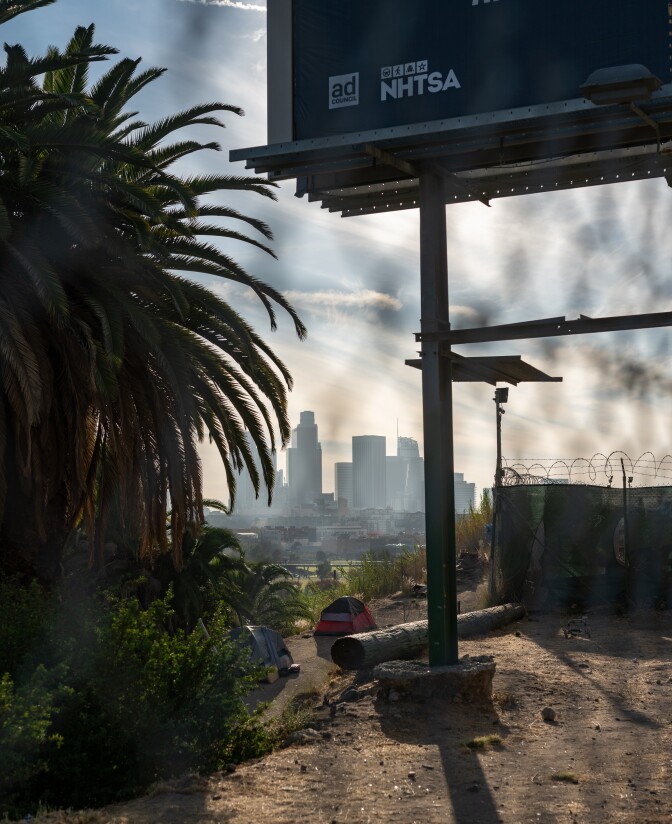 A photo of the downtown Los Angeles skyline taken from the view of the North Broadway bridge. There is a palm tree, tents and a billboard in the foreground. 