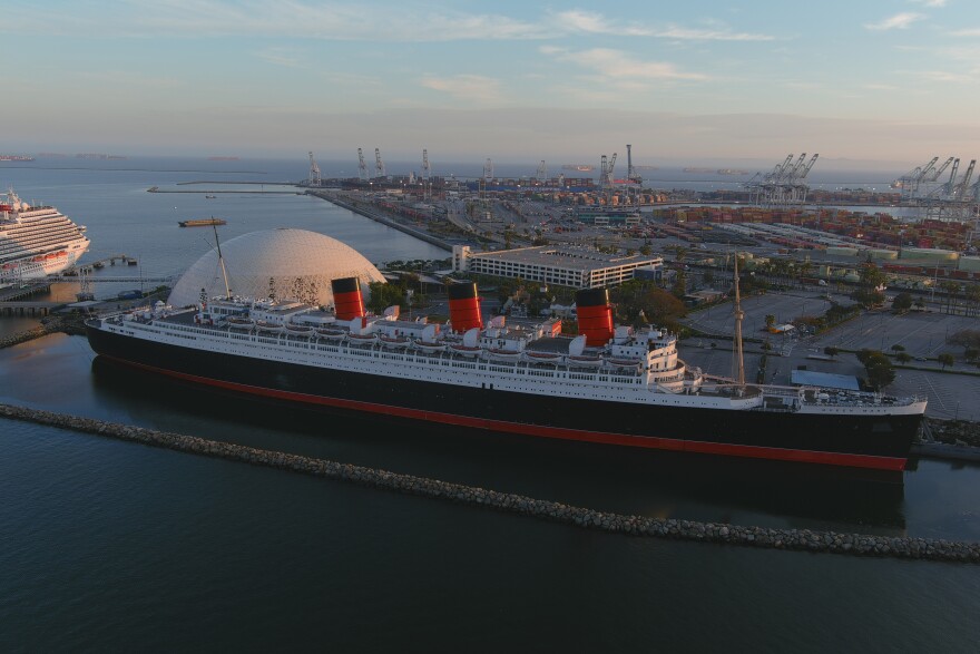 An aerial view of the Queen Mary has the ship in the foreground with it's well-known three red and black funnels. Behind the ship is a large white dome now used for cruise ship passengers.