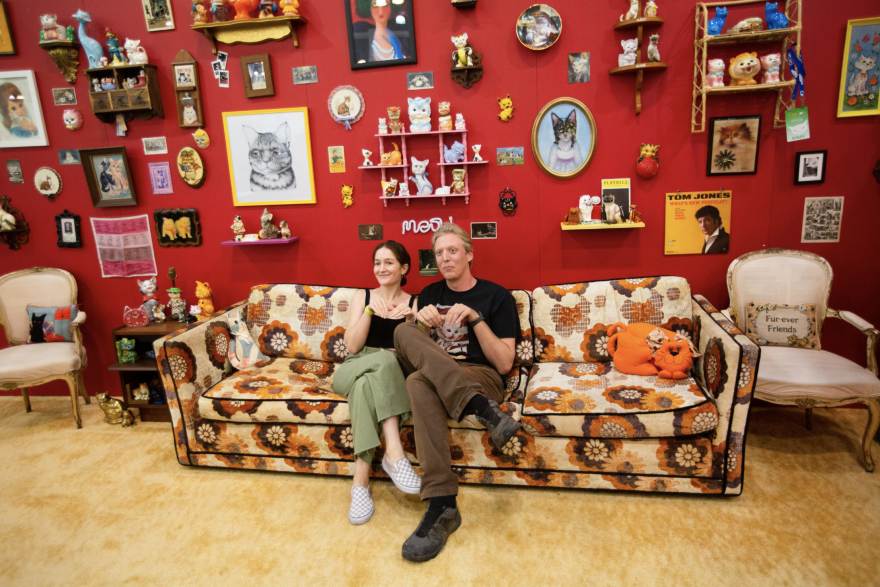 A man and a woman sitting on a coach in a living room. The wall behind them is decorated with cat pictures, photos and artworks.