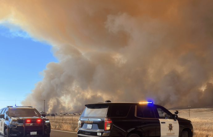 Two black and white law enforcement vehicles are on a road with dark plumes of smoke behind them. 