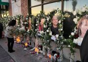 Large photos rest on easels rimming in white flowers with candles at the base. One women is bent over reading the messages.