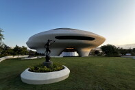 A saucer-shaped building sits on a lawn behind a bronze statue of a figure holding a pole atop a pedestal.