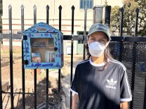A woman wearing a muted green baseball cap, a white face mask, and black t-shirt is looking ahead into the camera. She's standing next to a blue, purple, and pink wooden box affixed to a black metal gate in front of a several story tan building.