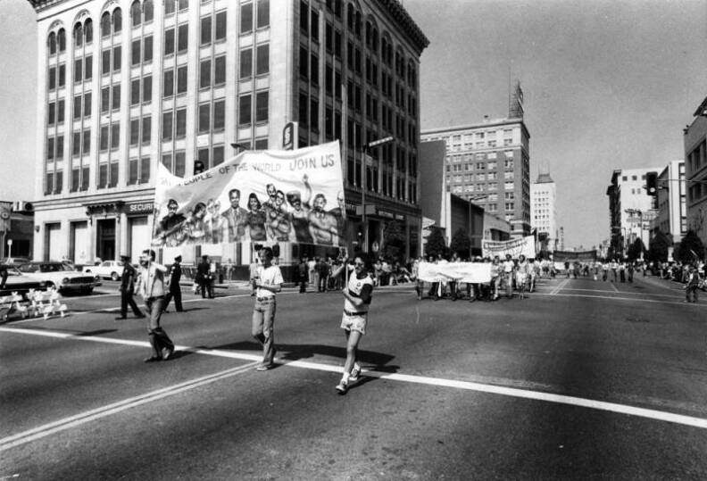A black and white wide view of people marching down a street. Three people are carrying a banner that reads Gay people of the World Join Us. The shot extends bak so you can see people marching in the distance.