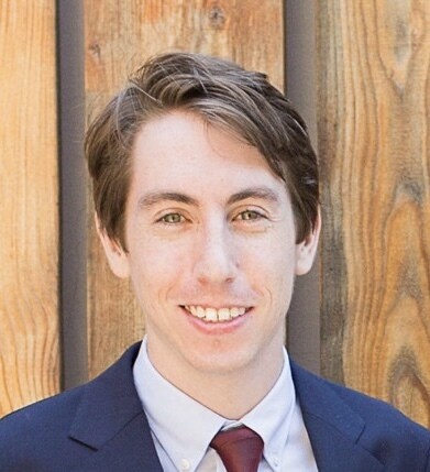 An image of a white man with a slight open smile, showing his teeth. He has sandy blond hair and is wearing a navy suit with a maroon tie, against a wooden backdrop.