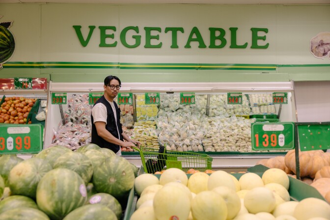 An Asian man with medium-light skin tone dark hair, black-rimmed glasses, wearing a black vest over a white shirt pushes a green shopping cart past a produce section in a super market. 