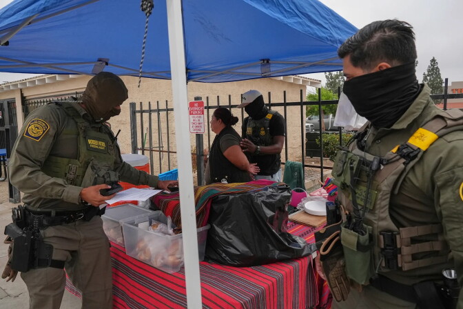 A woman standing underneath a blue pop up tent is detained by U.S. Border Patrol agents wearing army green uniforms. Underneath the tent are tables covered with red and grey striped tablecloths with various boxes and items.   